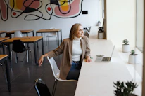 A woman is sitting on a chair in a modern room with tables and chairs. She is wearing a beige blazer over a white turtleneck and blue jeans, smiling and looking out the window. A laptop and a smartphone are placed on the desk in front of her, surrounded by potted plants. The background features a colorful abstract mural on the wall.