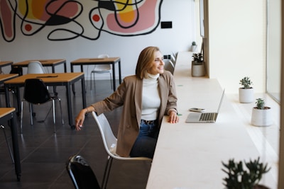 A woman is sitting on a chair in a modern room with tables and chairs. She is wearing a beige blazer over a white turtleneck and blue jeans, smiling and looking out the window. A laptop and a smartphone are placed on the desk in front of her, surrounded by potted plants. The background features a colorful abstract mural on the wall.