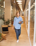 woman in blue dress shirt and blue denim jeans standing beside brown wooden chair