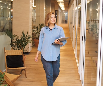 woman in blue dress shirt and blue denim jeans standing beside brown wooden chair