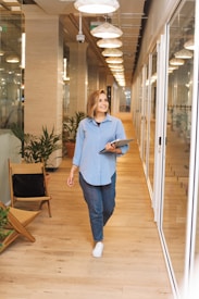 A person is walking down a bright, modern corridor with glass walls and wooden flooring. They are holding a laptop and smiling, dressed in casual attire. The corridor is decorated with potted plants and overhead lights, creating a welcoming office environment.