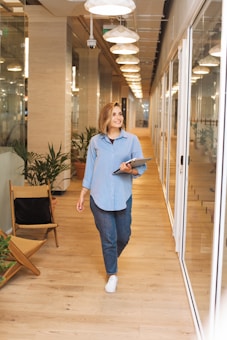 A person is walking down a bright, modern corridor with glass walls and wooden flooring. They are holding a laptop and smiling, dressed in casual attire. The corridor is decorated with potted plants and overhead lights, creating a welcoming office environment.