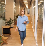 woman in blue dress shirt and blue denim jeans standing beside brown wooden chair