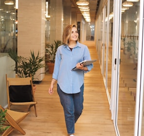 woman in blue dress shirt and blue denim jeans standing beside brown wooden chair