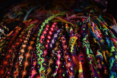 A vibrant array of intricately woven friendship bracelets displayed closely together. The bracelets feature a variety of patterns including zigzags, twists, and diamonds, showcasing a diverse palette of bright colors such as pinks, blues, greens, and yellows.