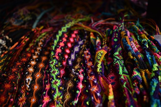 A vibrant array of intricately woven friendship bracelets displayed closely together. The bracelets feature a variety of patterns including zigzags, twists, and diamonds, showcasing a diverse palette of bright colors such as pinks, blues, greens, and yellows.