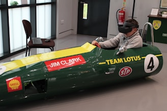 A person is seated inside a green vintage racing car simulator, wearing a virtual reality headset. The car displays 'Jim Clark' and 'Team Lotus' with various sponsor logos. A keyboard and mouse are placed on a nearby chair, and a fire extinguisher is visible on the wall.