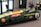 A person is seated inside a green vintage racing car simulator, wearing a virtual reality headset. The car displays 'Jim Clark' and 'Team Lotus' with various sponsor logos. A keyboard and mouse are placed on a nearby chair, and a fire extinguisher is visible on the wall.