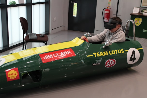 A person is seated inside a green vintage racing car simulator, wearing a virtual reality headset. The car displays 'Jim Clark' and 'Team Lotus' with various sponsor logos. A keyboard and mouse are placed on a nearby chair, and a fire extinguisher is visible on the wall.