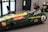 A person is seated inside a green vintage racing car simulator, wearing a virtual reality headset. The car displays 'Jim Clark' and 'Team Lotus' with various sponsor logos. A keyboard and mouse are placed on a nearby chair, and a fire extinguisher is visible on the wall.