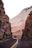A bus travels along a winding road flanked by tall, rugged cliffs. The cliffs display layers of red and white rock, showcasing impressive geological formations. Sparse vegetation can be seen clinging to the rock face, and the road is marked with a yellow double line down the center.