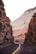 A bus travels along a winding road flanked by tall, rugged cliffs. The cliffs display layers of red and white rock, showcasing impressive geological formations. Sparse vegetation can be seen clinging to the rock face, and the road is marked with a yellow double line down the center.