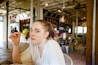 A woman with light hair is sitting in a casual, open restaurant setting. She is holding what appears to be a fry or small food item close to her mouth, looking directly at the camera. The interior features industrial-style decor with metal chairs and high ceilings. There are people in the background and large windows letting in natural light.