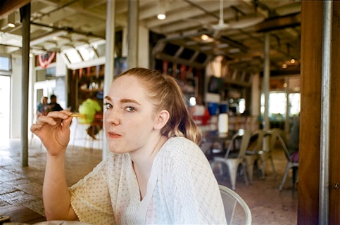 A woman with light hair is sitting in a casual, open restaurant setting. She is holding what appears to be a fry or small food item close to her mouth, looking directly at the camera. The interior features industrial-style decor with metal chairs and high ceilings. There are people in the background and large windows letting in natural light.