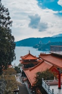 Traditional Chinese-style buildings with ornate red-tiled roofs are situated among lush green trees. A tranquil body of water and mountainous landscape can be seen in the distance under a partly cloudy sky.