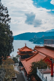 Traditional Chinese-style buildings with ornate red-tiled roofs are situated among lush green trees. A tranquil body of water and mountainous landscape can be seen in the distance under a partly cloudy sky.