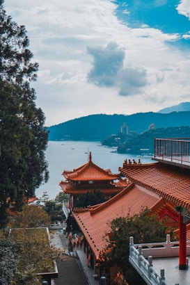 Traditional Chinese-style buildings with ornate red-tiled roofs are situated among lush green trees. A tranquil body of water and mountainous landscape can be seen in the distance under a partly cloudy sky.
