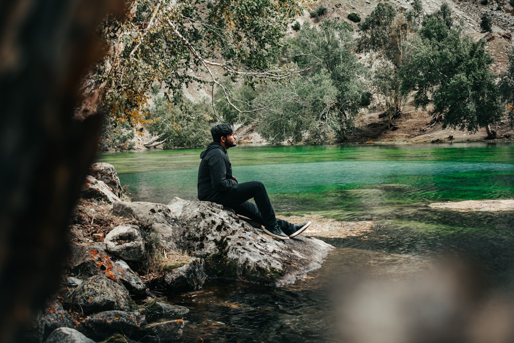 man in black jacket sitting on rock near river during daytime