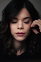 A woman with dark wavy hair appears relaxed with her eyes closed and a serene expression on her face. She gently touches her face with her right hand.