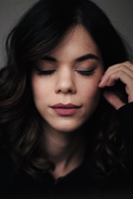 A woman with dark wavy hair appears relaxed with her eyes closed and a serene expression on her face. She gently touches her face with her right hand.