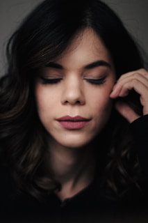 A woman with dark wavy hair appears relaxed with her eyes closed and a serene expression on her face. She gently touches her face with her right hand.