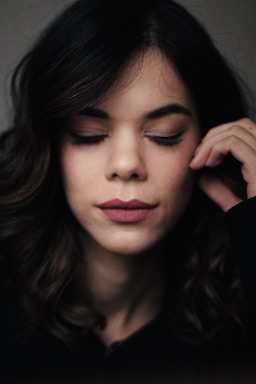 A woman with dark wavy hair appears relaxed with her eyes closed and a serene expression on her face. She gently touches her face with her right hand.