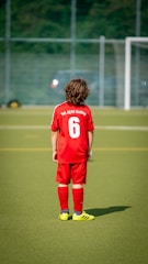 A young child is wearing a red soccer uniform with the number 6 on the back, standing on a green soccer field. The child is facing away towards a goalpost in the distance with a blurred background of a fence and trees.