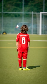 A young child is wearing a red soccer uniform with the number 6 on the back, standing on a green soccer field. The child is facing away towards a goalpost in the distance with a blurred background of a fence and trees.