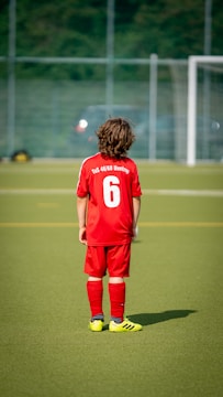 A young child is wearing a red soccer uniform with the number 6 on the back, standing on a green soccer field. The child is facing away towards a goalpost in the distance with a blurred background of a fence and trees.
