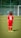 A young child is wearing a red soccer uniform with the number 6 on the back, standing on a green soccer field. The child is facing away towards a goalpost in the distance with a blurred background of a fence and trees.