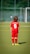 A young child is wearing a red soccer uniform with the number 6 on the back, standing on a green soccer field. The child is facing away towards a goalpost in the distance with a blurred background of a fence and trees.