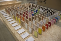 A display of colorful sample panels arranged in a grid on an OSB board table. Each sample is mounted on a wooden block with a variety of colors and textures. Labels with text descriptions are placed in front of the samples. In the background, there is a cardboard box with an IKEA logo.
