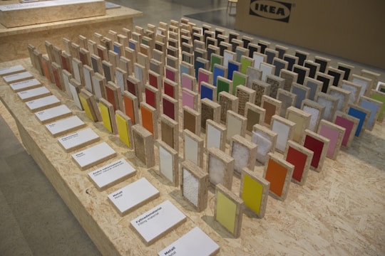 A display of colorful sample panels arranged in a grid on an OSB board table. Each sample is mounted on a wooden block with a variety of colors and textures. Labels with text descriptions are placed in front of the samples. In the background, there is a cardboard box with an IKEA logo.