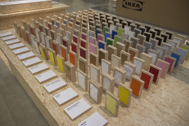 A display of colorful sample panels arranged in a grid on an OSB board table. Each sample is mounted on a wooden block with a variety of colors and textures. Labels with text descriptions are placed in front of the samples. In the background, there is a cardboard box with an IKEA logo.