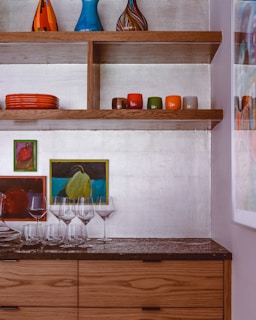 A modern kitchen or dining room area with wooden shelves displaying colorful vases on the top shelf. The second shelf holds several small, brightly colored bowls or cups. Below, there are two small paintings with vibrant colors and several clear wine glasses and tumblers neatly arranged on a dark countertop. The cabinets have a natural wood finish, complementing the overall aesthetic.