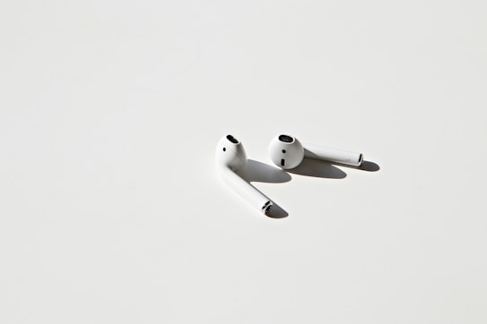A pair of white wireless earbuds placed on a light gray surface, casting soft shadows. The minimalistic design and clean lines are highlighted by the stark contrast between the earbuds and the background.