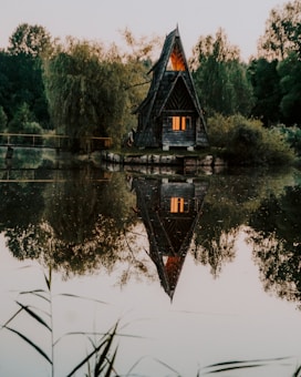 A rustic, triangular wooden cabin sits amidst lush greenery on a serene lake with its reflection mirroring on the water. The cabin emits a warm glow from its windows, adding a cozy contrast to the cool hues of the surrounding trees and water.