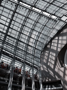A large, modern glass atrium with a grid-like roof structure, allowing natural light to cast geometric shadows on the walls and floors. The space is expansive and open, with a row of international flags hanging from the upper level.