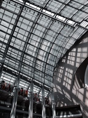 A large, modern glass atrium with a grid-like roof structure, allowing natural light to cast geometric shadows on the walls and floors. The space is expansive and open, with a row of international flags hanging from the upper level.