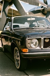 A vintage black car with a chrome grille and round headlights is parked on a concrete surface. The car is positioned in front of a large aircraft propeller, hinting at an aviation setting. The sunlight casts shadows on the car's surface, highlighting its classic contours and shiny paint.
