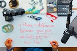 A workspace setup featuring a variety of objects such as a camera, a notepad with colorful artwork, a sound mixer, a microphone, glasses, pens, and a drink. In the center, a whiteboard contains a hand-written question in red ink asking about the difference between personal and divine plans.