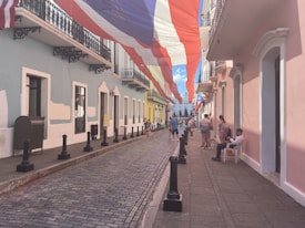 A narrow, cobblestone street lined with colorful colonial-style buildings under a large red, white, and blue flag. People are gathered on the sidewalk, some sitting and others standing. Wrought iron balconies and pastel-painted walls add to the charm of the street.