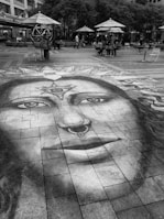 A large, detailed chalk drawing of a face with symbolic designs is created on a paved urban plaza. The drawing features unique details like a nose ring and patterns on the forehead. It extends across the tiles, attracting attention. Surrounding the artwork, people are sitting or walking under large umbrellas and trees, giving the space a casual, social atmosphere.