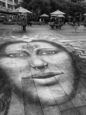A large, detailed chalk drawing of a face with symbolic designs is created on a paved urban plaza. The drawing features unique details like a nose ring and patterns on the forehead. It extends across the tiles, attracting attention. Surrounding the artwork, people are sitting or walking under large umbrellas and trees, giving the space a casual, social atmosphere.