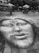 A large, detailed chalk drawing of a face with symbolic designs is created on a paved urban plaza. The drawing features unique details like a nose ring and patterns on the forehead. It extends across the tiles, attracting attention. Surrounding the artwork, people are sitting or walking under large umbrellas and trees, giving the space a casual, social atmosphere.