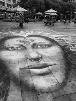 A large, detailed chalk drawing of a face with symbolic designs is created on a paved urban plaza. The drawing features unique details like a nose ring and patterns on the forehead. It extends across the tiles, attracting attention. Surrounding the artwork, people are sitting or walking under large umbrellas and trees, giving the space a casual, social atmosphere.