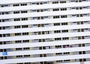 A tall residential building with multiple rows of uniform balconies. Each balcony is enclosed with rails, and some have laundry or small items hanging. The facade is primarily white with consistent horizontal lines formed by the balcony railings.