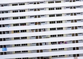 A tall residential building with multiple rows of uniform balconies. Each balcony is enclosed with rails, and some have laundry or small items hanging. The facade is primarily white with consistent horizontal lines formed by the balcony railings.