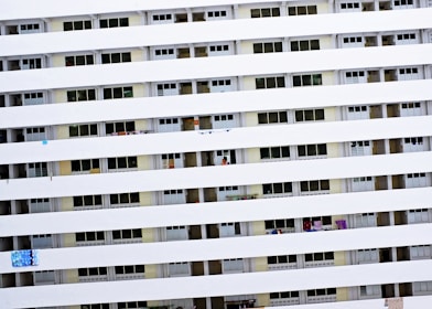A tall residential building with multiple rows of uniform balconies. Each balcony is enclosed with rails, and some have laundry or small items hanging. The facade is primarily white with consistent horizontal lines formed by the balcony railings.
