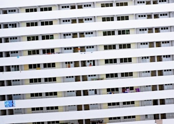 A tall residential building with multiple rows of uniform balconies. Each balcony is enclosed with rails, and some have laundry or small items hanging. The facade is primarily white with consistent horizontal lines formed by the balcony railings.